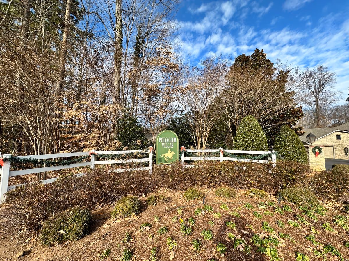 Landscaped entrance with a green 'Polo Club Villas' sign in front of a white fence, shrubs, and trees under a blue sky.