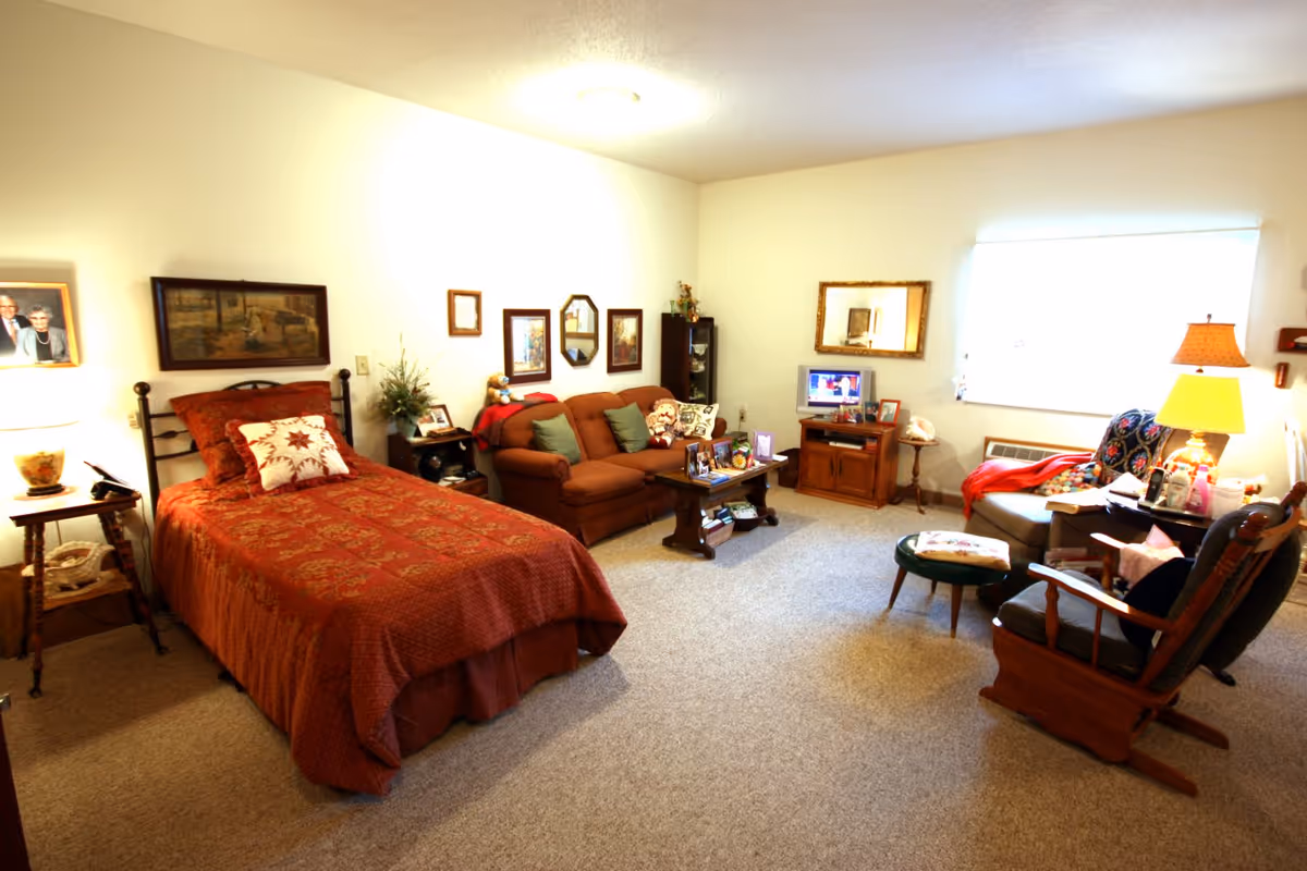 A cozy senior living room with a single bed covered in a red patterned quilt, a small brown couch with green pillows, a wooden coffee table with framed photos, a TV on a wooden stand, and two armchairs near a window with a lamp on a side table. The walls are decorated with framed pictures and mirrors.