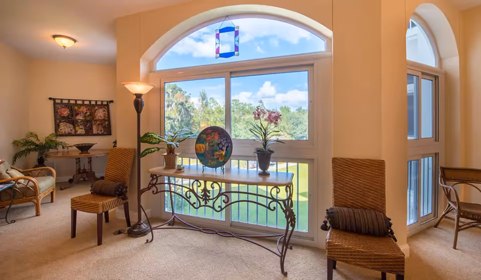 A bright and cozy interior room with large arched windows showing a view of green trees and blue sky. The room features a decorative table with potted plants and a colorful plate, two wicker chairs with cushions, a floor lamp, and a small table with a plant and decorative items in the background.