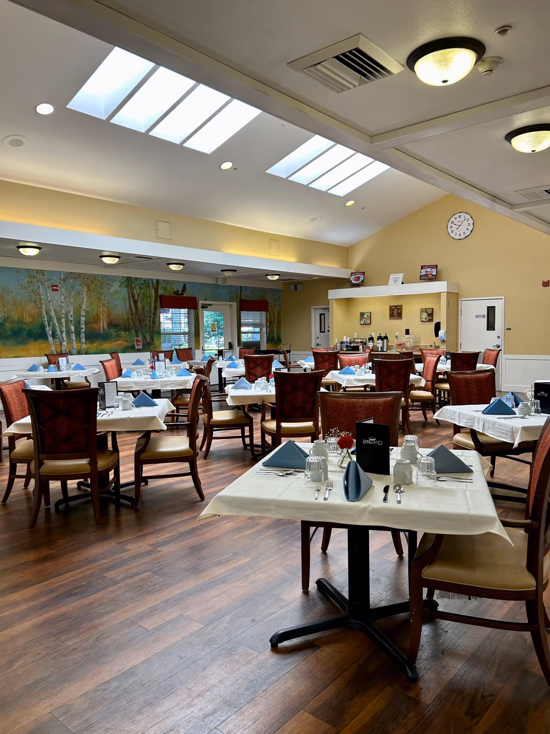 A spacious dining room with multiple tables covered in white tablecloths, each set with blue folded napkins, glasses, and silverware. The room has wooden floors, a mural of trees on one wall, skylights on the ceiling, and warm lighting fixtures. There is a clock on the far wall and a beverage station in the back.