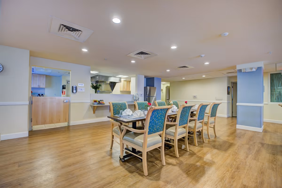 A spacious dining room in a senior living facility with a long table set for a meal. The table has eight chairs with blue patterned upholstery and cream-colored cushions. The room has wood flooring, recessed ceiling lights, and a view into a kitchen area with stainless steel appliances and wooden cabinets.