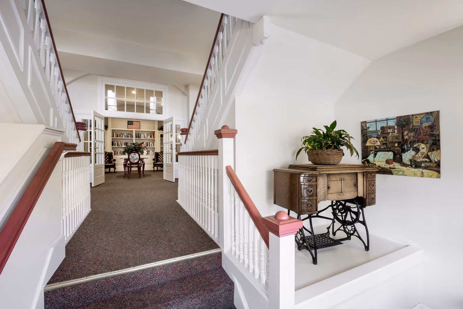 Carpeted split staircase and hallway leading to a library with bookshelves, with an antique sewing table and potted plant displayed on the right.