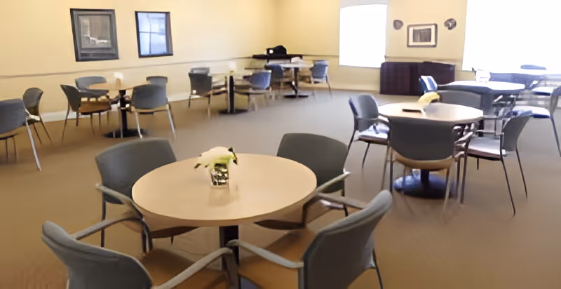 A spacious dining room with multiple round tables and chairs arranged around them. The room has beige walls with framed pictures and a large window letting in natural light. A small flower arrangement is placed on the closest table.