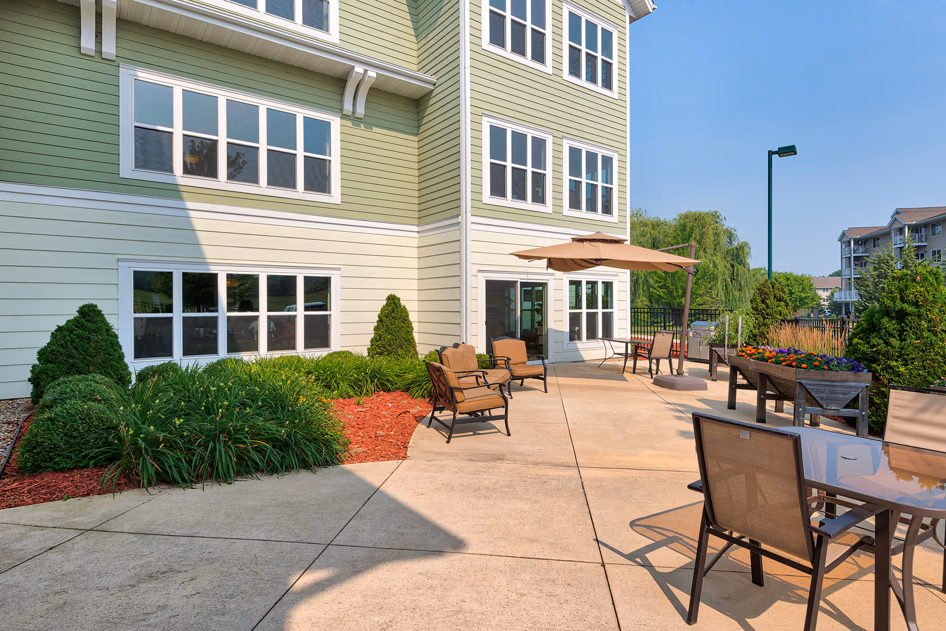Outdoor patio area at a senior living facility with several cushioned chairs, a table with an umbrella, raised garden beds with flowers, and a green and beige building exterior under a clear blue sky.
