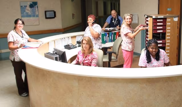A group of six healthcare workers in a nursing facility reception area. They are wearing scrubs and are engaged in various activities such as talking on the phone, working on computers, and organizing files behind a curved reception desk.