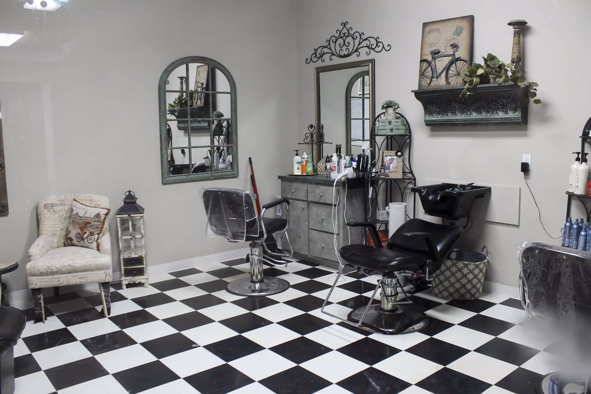Interior of a hair salon area with black and white checkered floor, two salon chairs, a washing station, a cushioned armchair with a decorative pillow, a tall lantern, mirrors on the walls, and shelves with hair care products and decorative items.
