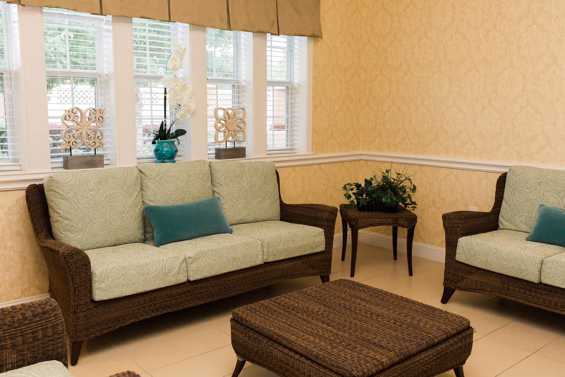 A cozy sitting area with wicker furniture including a sofa, armchair, and ottoman. The sofa and armchair have light green patterned cushions with teal accent pillows. Behind the sofa are three windows with white blinds and decorative items on the windowsill, including a turquoise vase with white flowers and two ornamental pieces. A small wicker side table with a green plant is placed in the corner. The walls are covered with light yellow patterned wallpaper and a white chair rail molding.