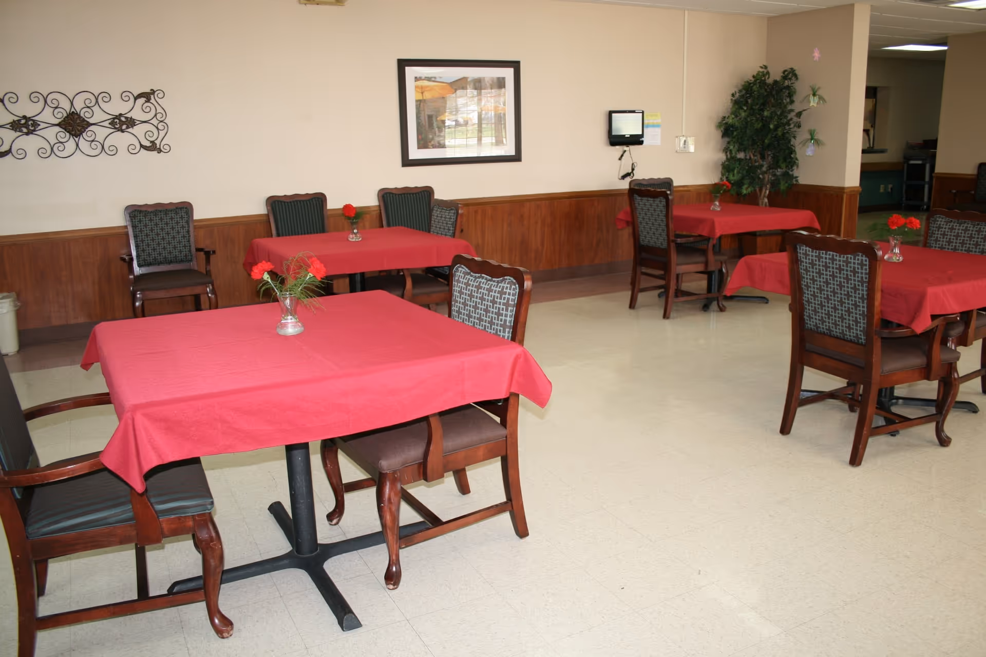 Dining area with multiple tables draped in red tablecloths, wooden chairs, and small vases holding red flowers.