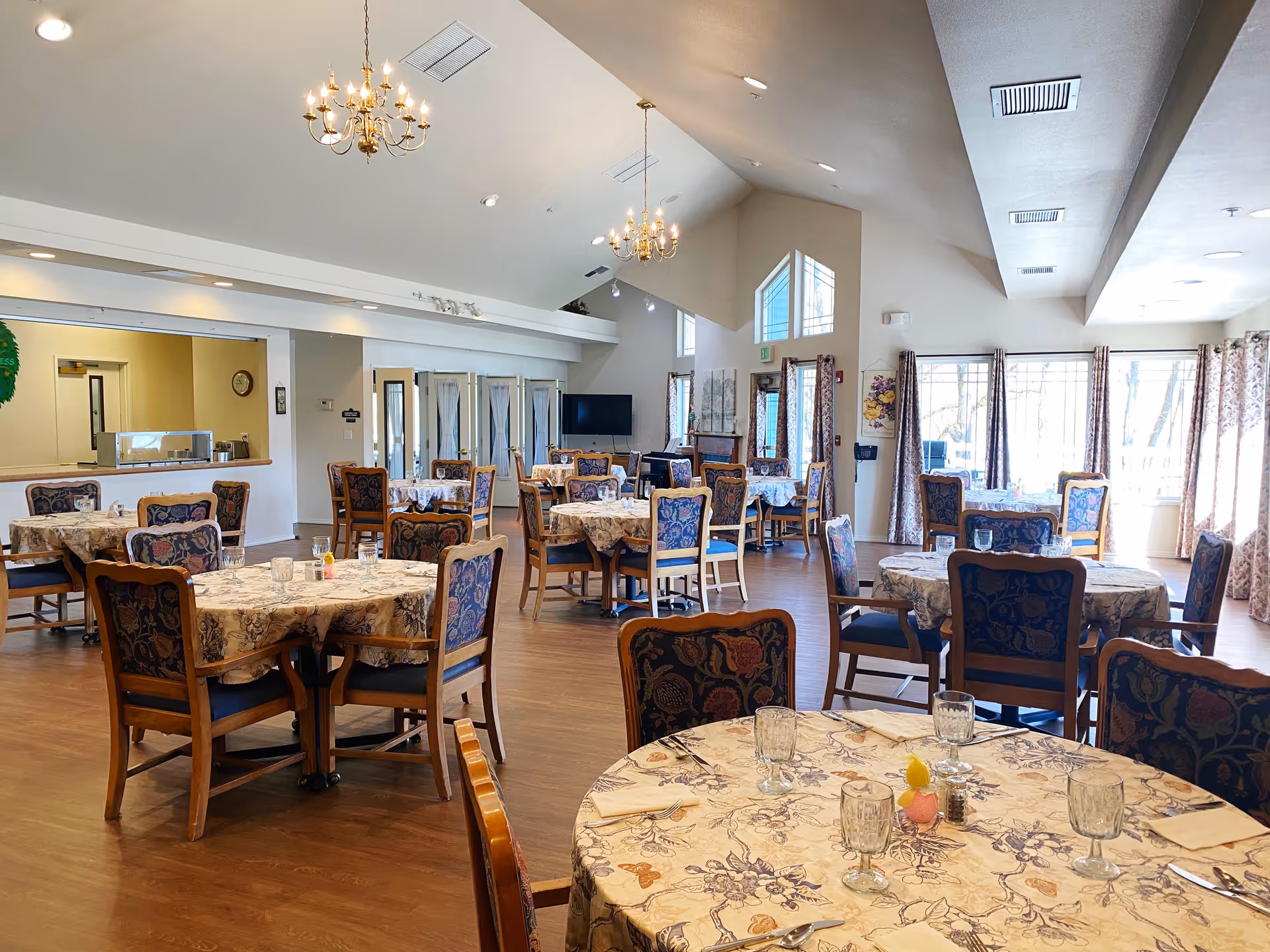 A spacious dining room in a retirement and assisted living facility with multiple round tables covered with floral tablecloths. Each table is set with glasses, napkins, and cutlery. The room has wooden chairs with patterned upholstery, large windows with curtains allowing natural light, and two chandeliers hanging from the ceiling. There is a TV mounted on the far wall and a serving counter on the left side.