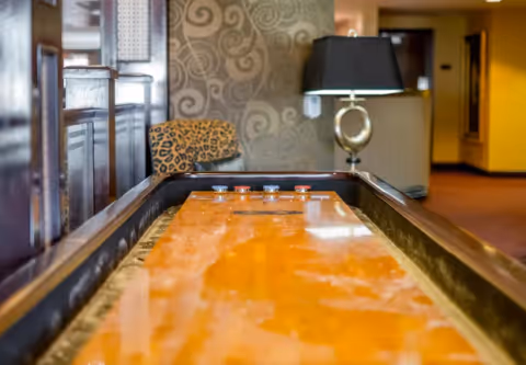 A shuffleboard table in a cozy indoor setting with a leopard print chair and a decorative lamp in the background, featuring warm lighting and patterned wallpaper.