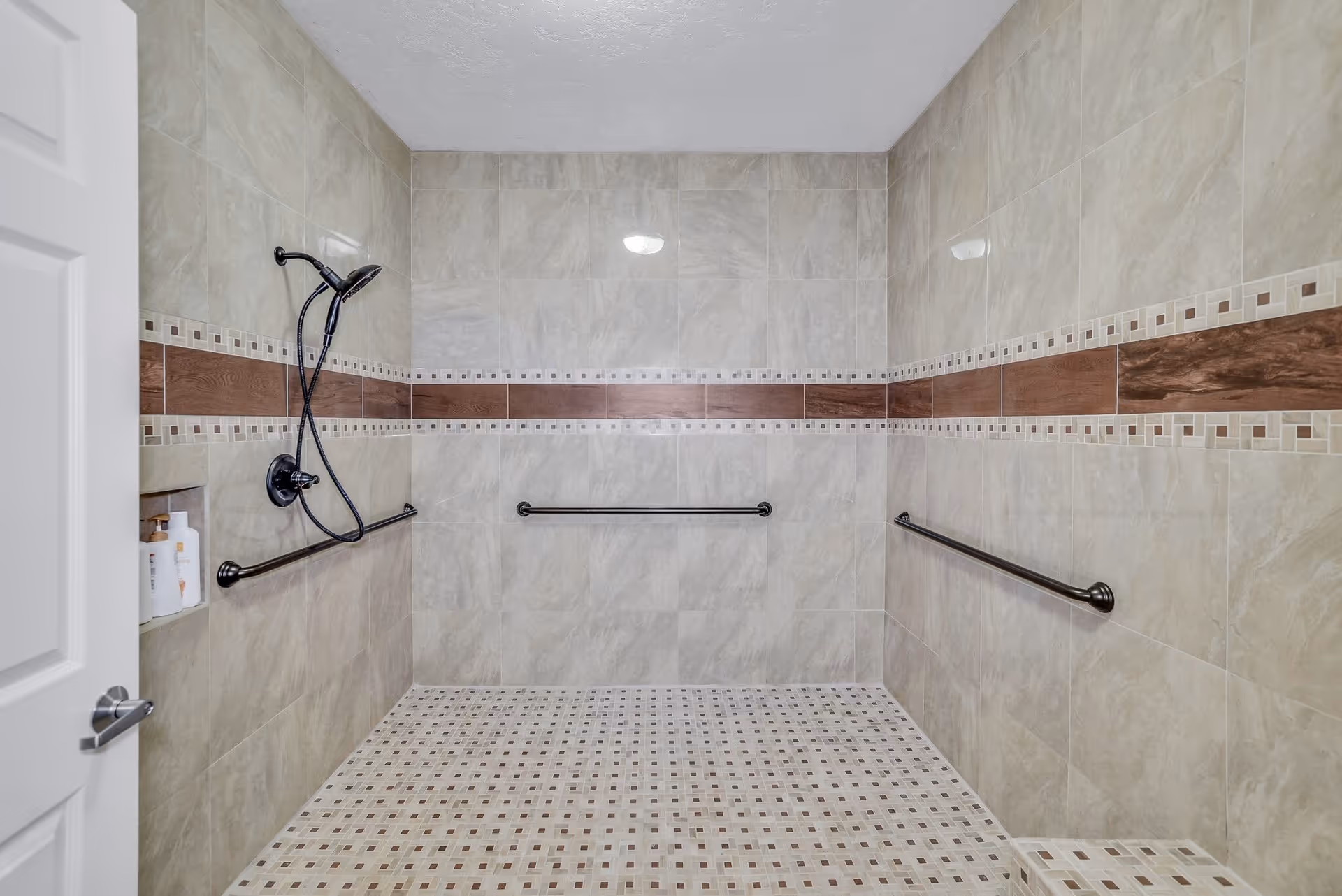 A spacious walk-in shower with beige and brown tiled walls and floor. The shower features a handheld showerhead, three black grab bars for support, and a built-in niche holding bottles of shampoo and soap. The door to the shower is partially open.