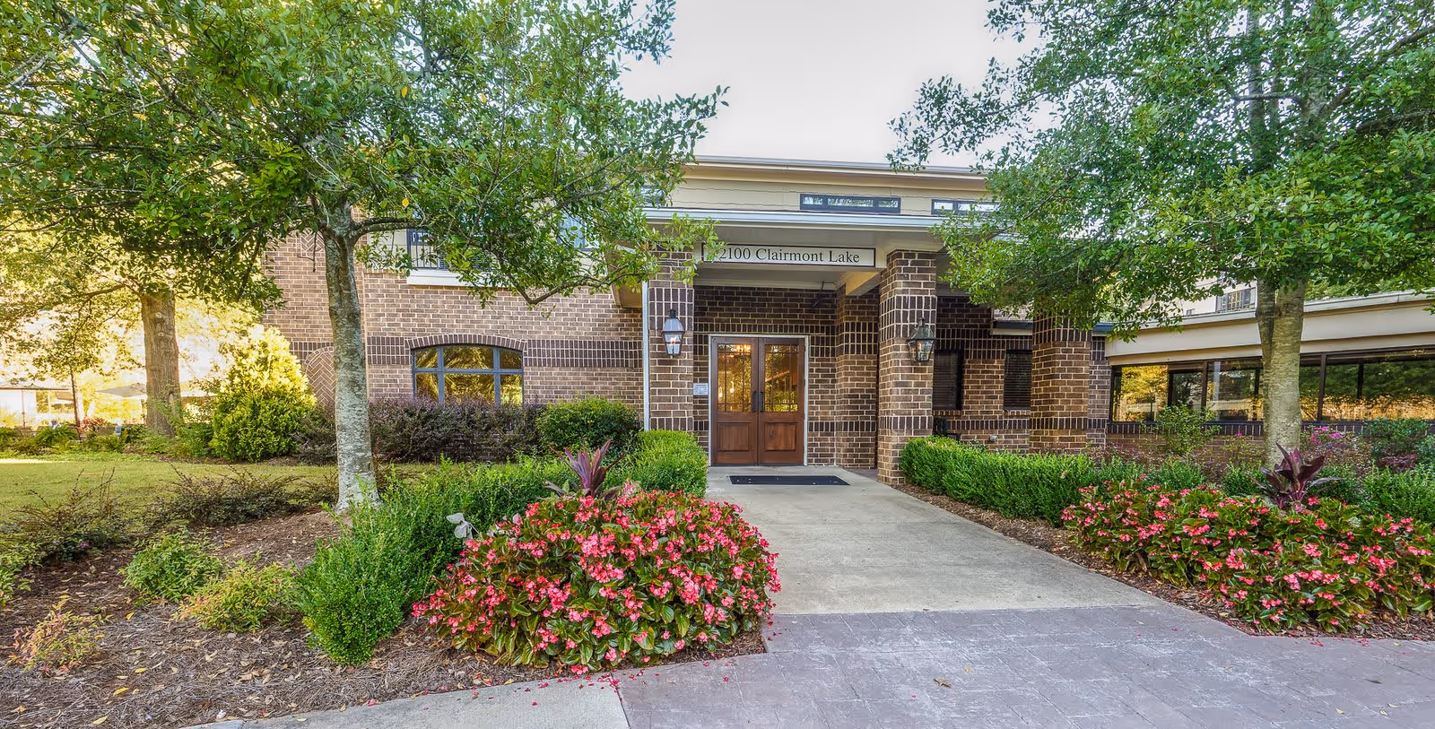 Front exterior view of a brick building with double wooden doors under a covered entrance. The building is surrounded by well-maintained landscaping including green bushes, pink flowers, and trees. A concrete pathway leads to the entrance.