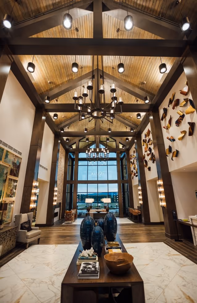 High-ceilinged lobby interior with chandeliers, floor-to-ceiling windows, seating areas, and a central table topped with decorative vases.
