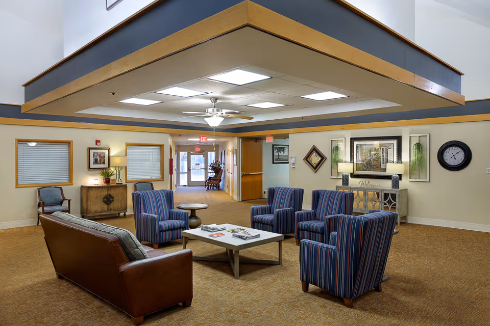 Lobby-style common room with a leather sofa, striped armchairs, coffee table, and console table in a senior living facility.