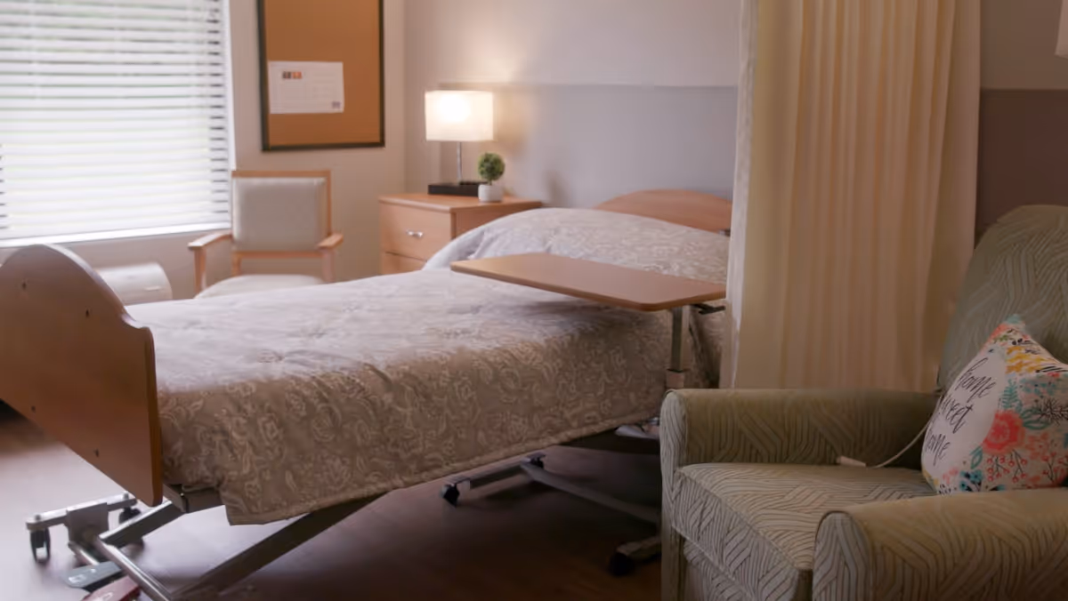 A nursing facility room with a hospital bed covered in a patterned beige bedspread, a small overbed table, a green armchair with a floral pillow that says 'Home Sweet Home', a wooden nightstand with a lamp and a small plant, a window with blinds, and a wooden chair next to the window.