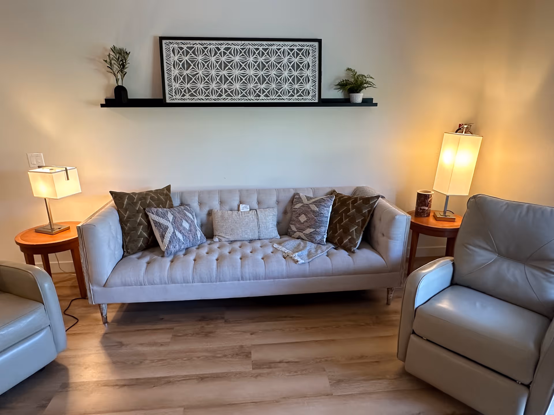 A cozy living room with a light gray tufted sofa adorned with five decorative pillows and a throw blanket. On either side of the sofa are wooden side tables, each with a modern lamp. Two gray leather armchairs are partially visible on the left and right sides. Above the sofa is a black floating shelf holding a framed geometric artwork and two small potted plants. The room has light wood flooring and neutral-colored walls.