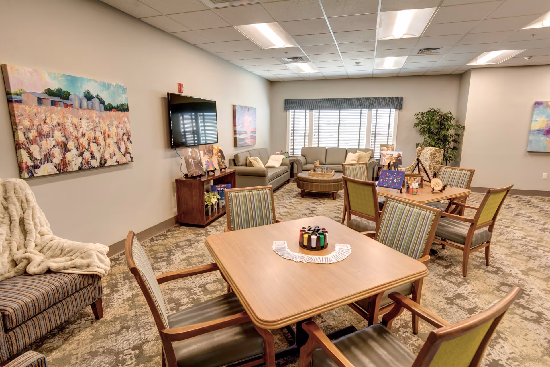 A cozy senior living common area with several wooden tables and chairs arranged for group activities. The room features a large window with blinds, a flat-screen TV mounted on the wall, comfortable sofas, and armchairs. There are paintings on the walls and a potted plant in the corner, creating a warm and inviting atmosphere.