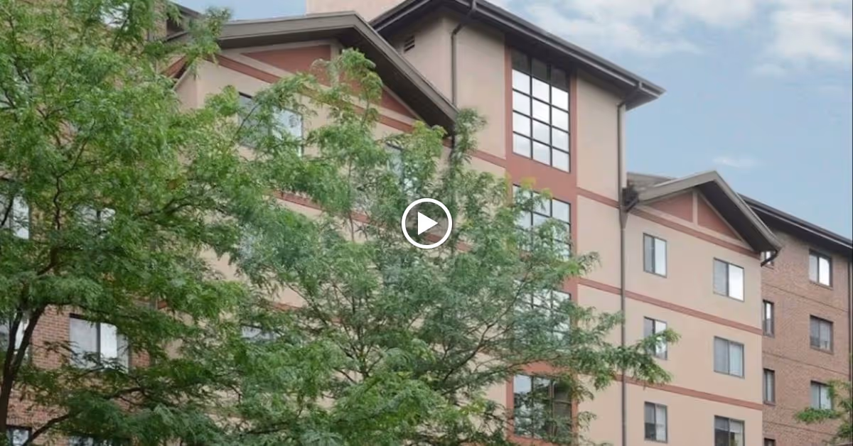 Exterior view of a multi-story senior living facility building partially obscured by green leafy trees under a blue sky.