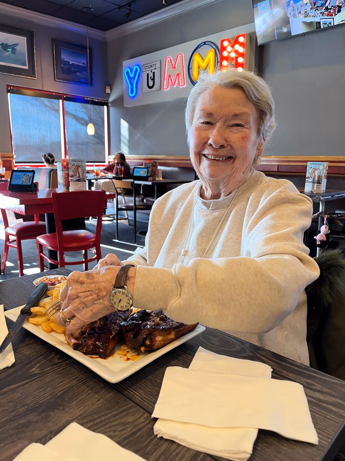 An elderly woman smiling and sitting at a restaurant table with a plate of ribs and fries in front of her. The restaurant interior has red chairs, tables, and a neon sign on the wall that spells 'YUMMY'. Other diners are visible in the background.