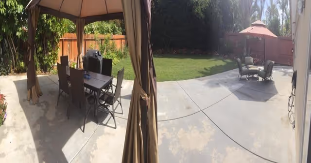 Outdoor patio area with two seating arrangements under umbrellas. One area has a table with six chairs under a gazebo, and the other has a smaller table with four chairs under a separate umbrella. The patio is surrounded by a wooden fence and greenery including trees and bushes.