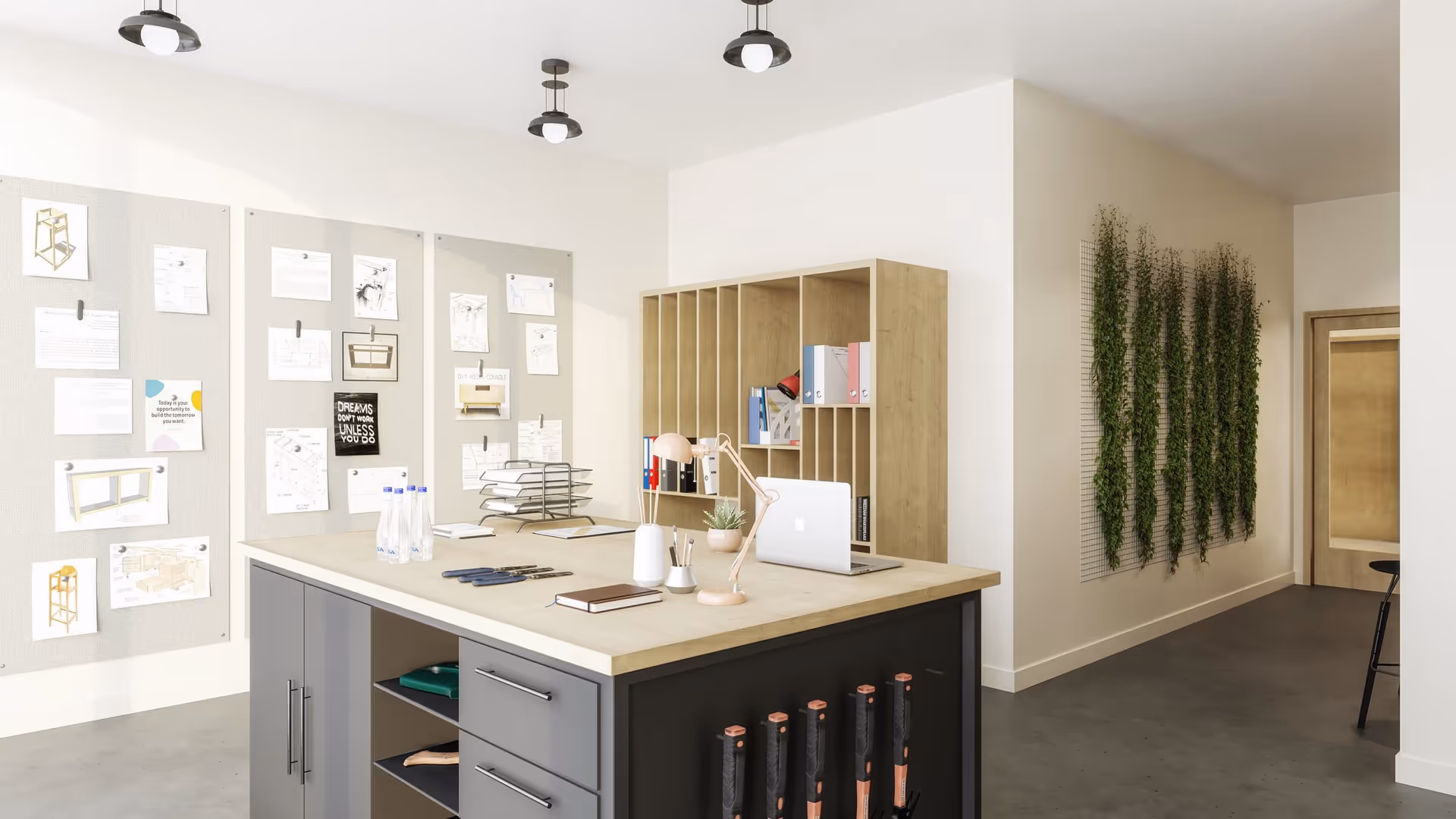 A modern office or workspace with a large wooden table in the center holding a laptop, desk lamp, stationery, and water bottles. Behind the table is a wooden shelving unit with binders and books. On the left wall, there are several pinned papers and sketches. The right wall features a vertical garden with green plants growing on a trellis. The floor is concrete, and the space is well-lit with ceiling lights.