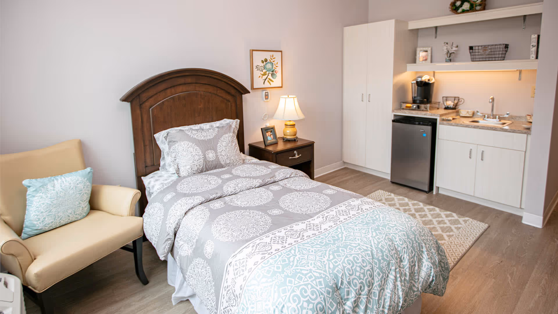 A furnished studio-style bedroom with a single bed, nightstand and lamp, an armchair, and a small kitchenette with sink and mini-fridge.