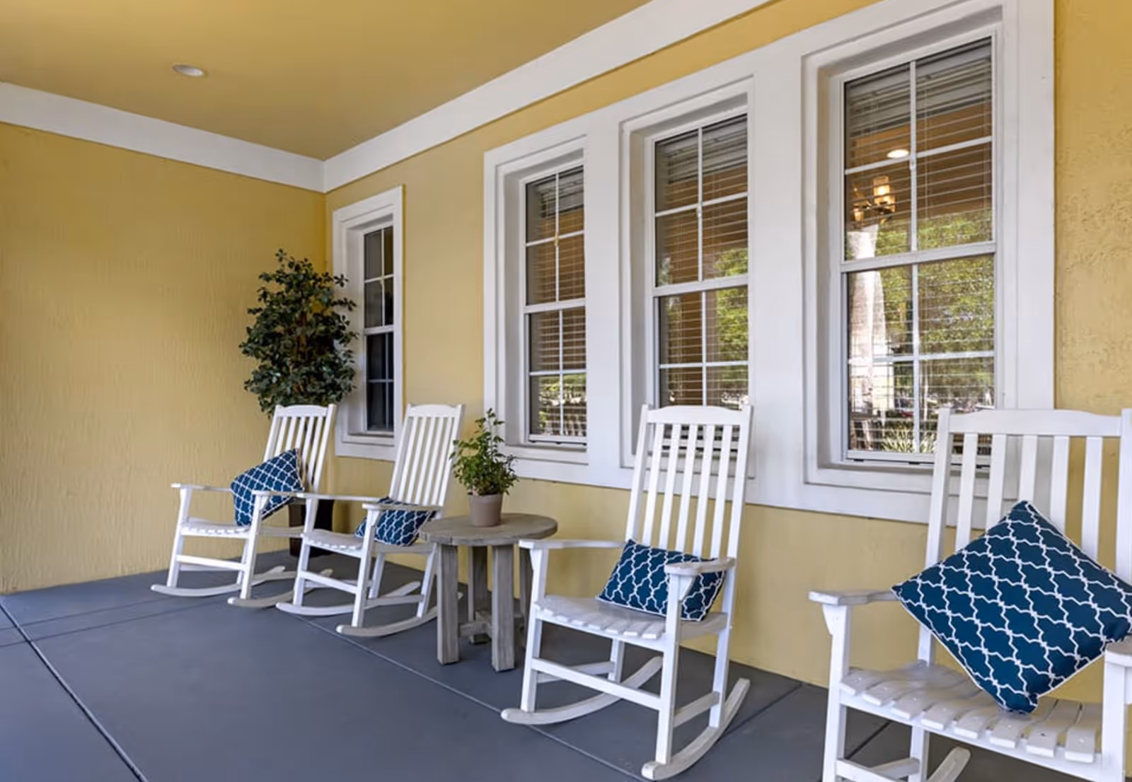 A covered porch area with four white wooden rocking chairs, each with a blue patterned cushion. There is a small round wooden table with a potted plant between two of the chairs. The wall behind the chairs is painted yellow and has three white-framed windows with blinds. A tall potted plant is placed in the corner.