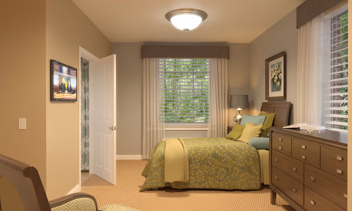 A cozy bedroom with a bed covered in green and yellow patterned bedding, multiple pillows, a wooden dresser with an open book and glasses on top, a window with white blinds and curtains, a bedside table with a lamp, a chair in the foreground, and a wall-mounted TV showing a news broadcast. The door to an adjacent bathroom is open, revealing a shower curtain with a floral pattern.
