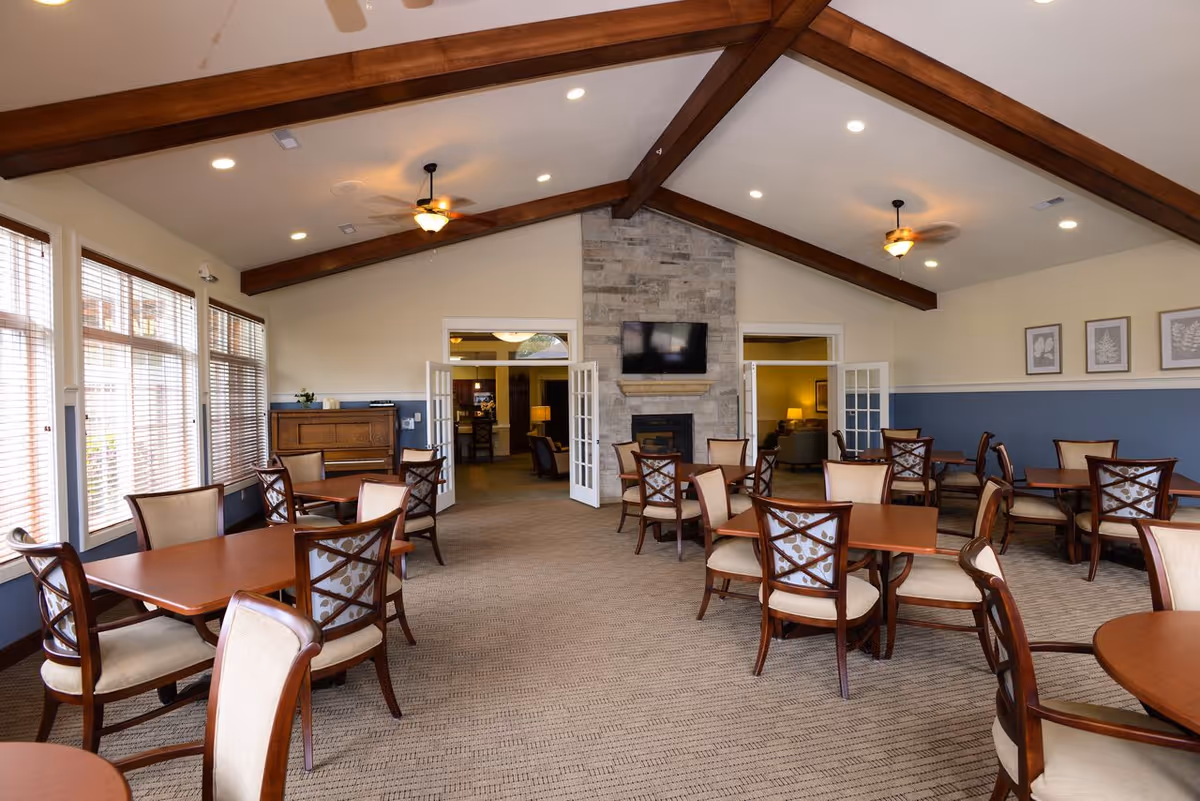 A spacious dining room with multiple wooden tables and cushioned chairs arranged neatly. The room features large windows with blinds on the left side, a stone fireplace with a mounted flat-screen TV in the center, and a high ceiling with exposed wooden beams and ceiling fans. The walls are painted in a two-tone style with blue on the lower half and cream on the upper half, decorated with framed artwork.