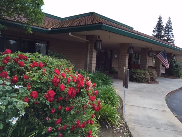 Exterior view of Legacy Oaks of Sacramento facility showing a covered entrance with hanging lanterns, a curved driveway, an American flag, and vibrant red roses and green shrubs in the foreground.