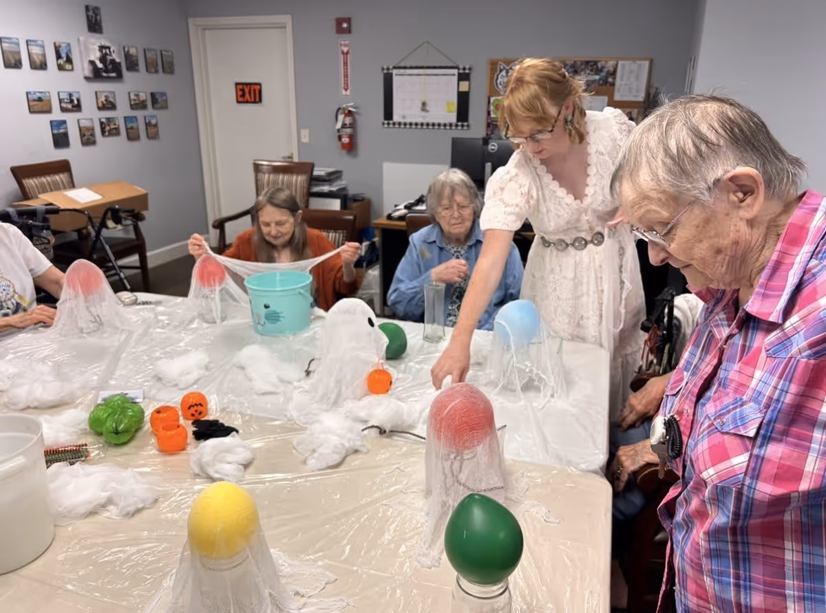 A group of elderly women and a younger woman are gathered around a table in a room, engaging in a craft activity involving balloons covered with white mesh and cotton to create ghost decorations. The room has chairs, a door with an exit sign, a fire extinguisher, and pictures on the wall.