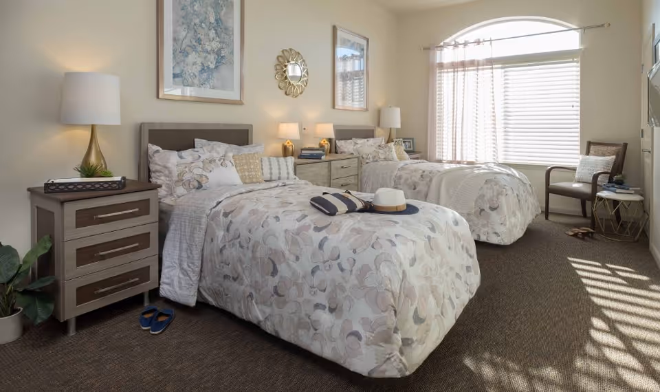 A bright and cozy bedroom with two twin beds featuring floral patterned bedding. Each bed has multiple pillows and a bedside table with lamps. There is a window with blinds and sheer curtains allowing natural light to fill the room. A chair with a cushion and a small round side table are placed near the window. The room is decorated with framed artwork and a decorative mirror on the wall.