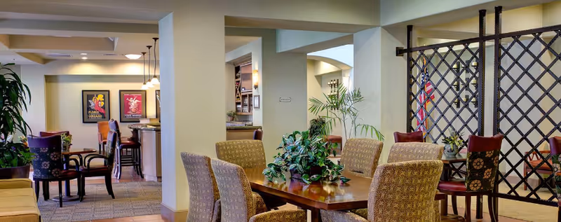 A spacious and well-lit common area in a senior living facility featuring multiple seating arrangements with upholstered chairs around wooden tables. There are decorative plants on the tables and in the corners, framed artwork on the walls, and a lattice partition with an American flag in the background.