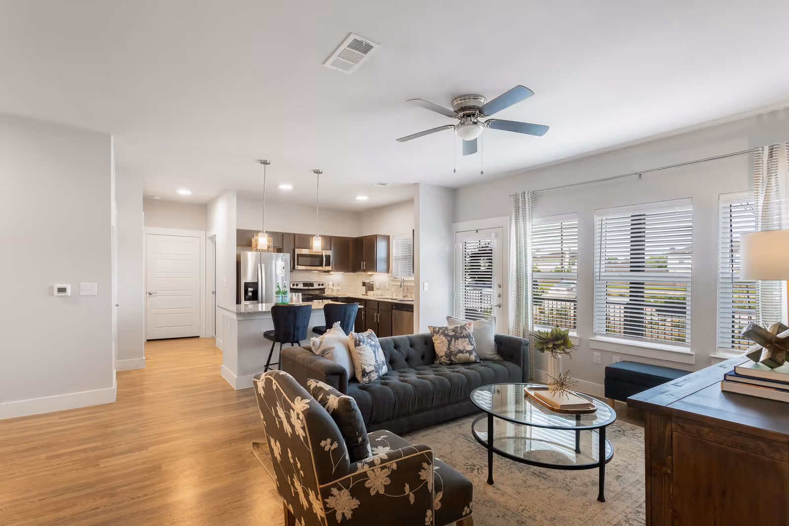 A bright and modern living room and kitchen area with wooden flooring. The living room features a dark gray tufted sofa with floral and plain cushions, a floral patterned armchair, a glass coffee table with decorative items, and a ceiling fan. The kitchen has dark wood cabinets, stainless steel appliances including a refrigerator, stove, and microwave, and two pendant lights hanging over a breakfast bar with two dark blue bar stools. Large windows with white blinds allow natural light to fill the space.
