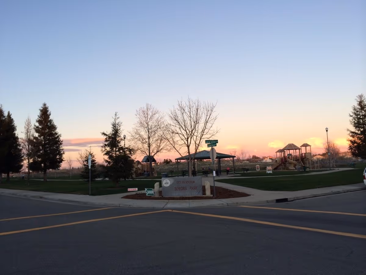 A park at sunset with a playground, trees, a covered picnic area, and a sign that reads 'City of Rocklin Sonora Park'. The sky is clear with a gradient from blue to orange near the horizon.