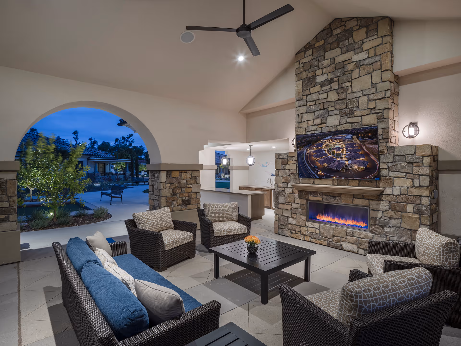 Covered outdoor lounge with wicker sofas and chairs arranged around a coffee table in front of a stone fireplace with a mounted TV, opening onto a landscaped courtyard.