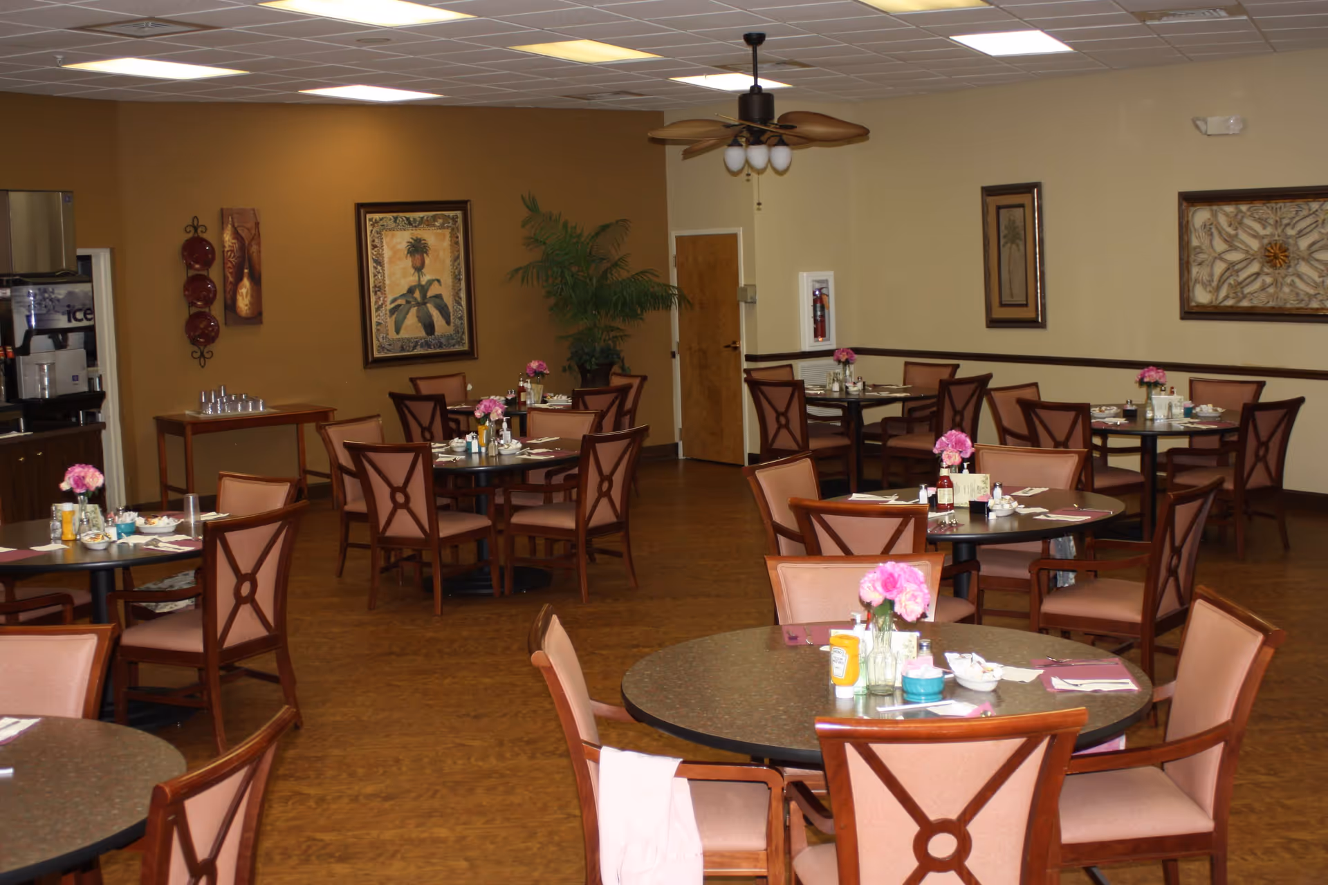 Dining room with multiple round tables and chairs arranged neatly. Each table has pink flower centerpieces, condiments, and place settings. The room has beige and brown walls decorated with framed artwork and a ceiling fan with lights. A beverage station is visible on the left side.