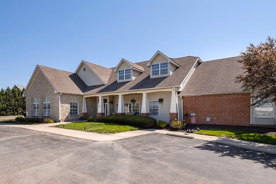 Exterior view of a single-story senior living facility building with a combination of stone and brick facade, a covered porch with hanging flower baskets, and a paved driveway in front under a clear blue sky.