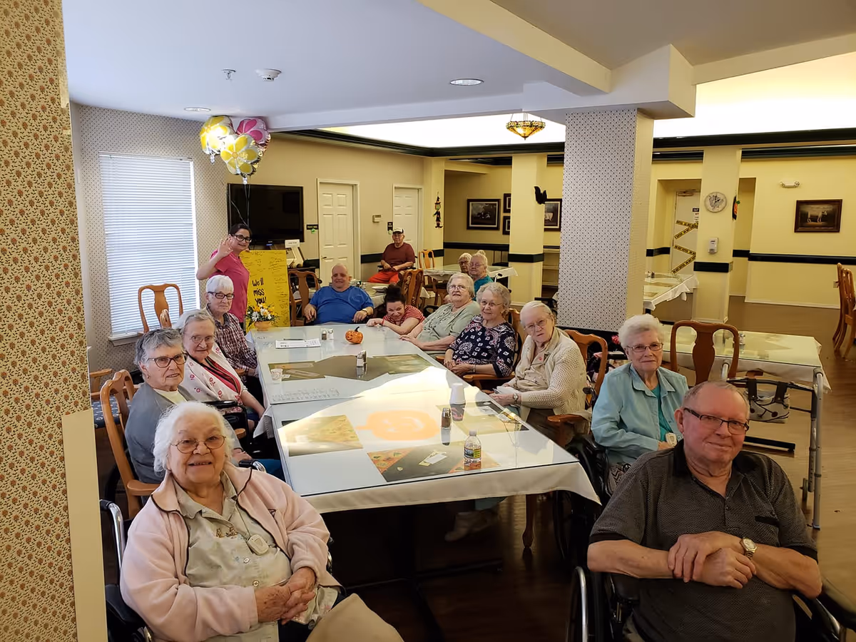 A group of elderly people sitting around a large dining table in a well-lit room with yellow walls and patterned wallpaper. A staff member stands near the window holding balloons and a sign that says 'We'll miss you!'. The room has wooden chairs and tables, and some decorations on the walls.