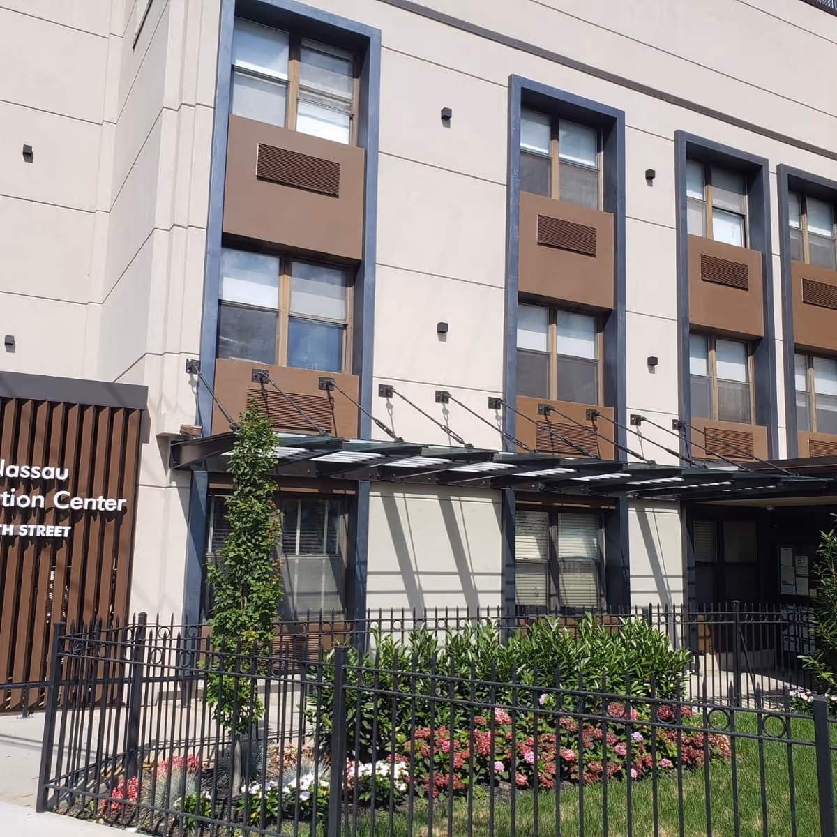 Exterior front of a multi-story rehabilitation and nursing center building with windows, a metal awning, landscaping and a black fence.