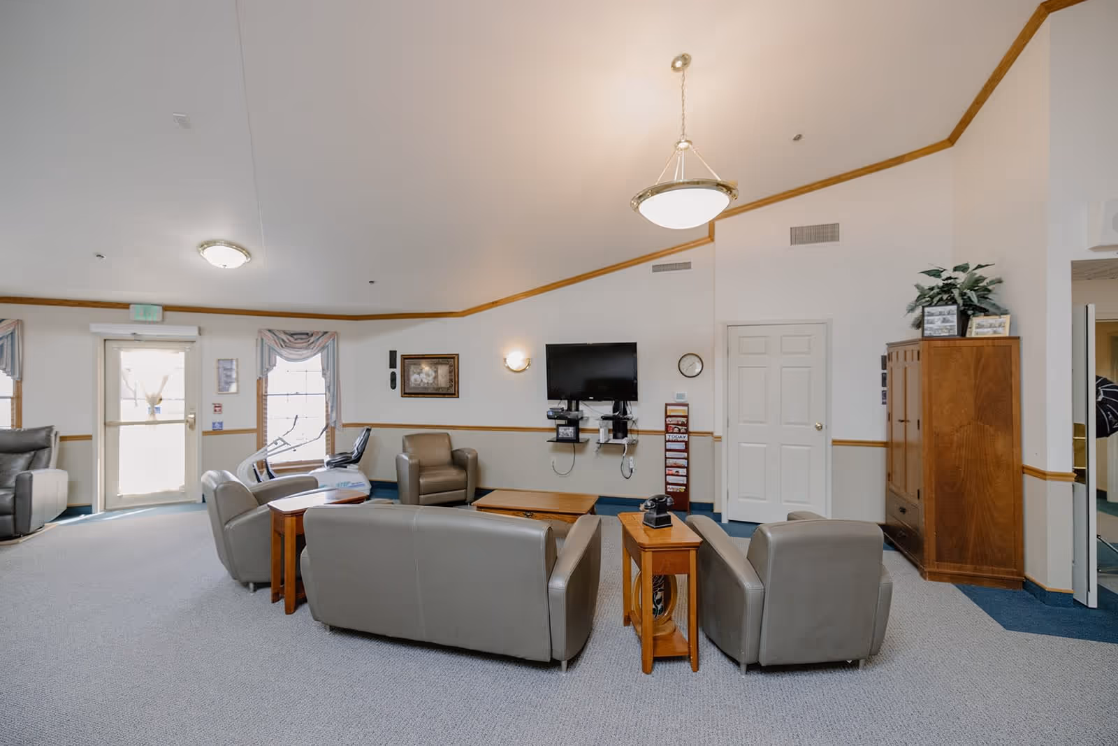 A spacious assisted living facility common area with beige leather sofas and armchairs arranged around wooden coffee and side tables. A flat-screen TV is mounted on the wall above a small shelf, with a clock and a brochure stand nearby. The room has light-colored walls with wooden trim, carpeted floors, two windows with curtains, and a door leading outside. A wooden cabinet with framed photos and a plant is visible on the right side.