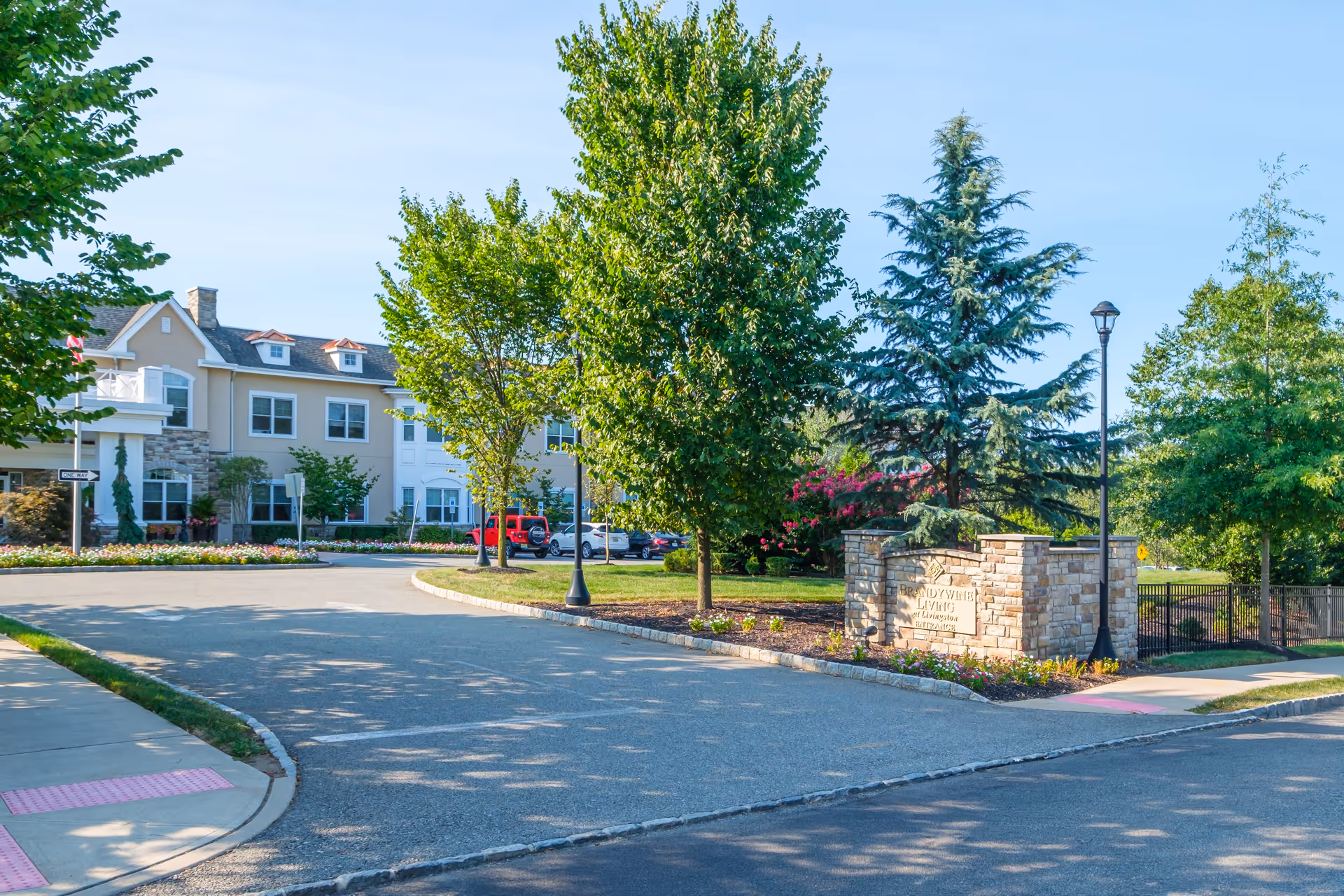 Entrance driveway to Brandywine Livingston by Monarch facility with a stone sign, trees, landscaped flower beds, and a two-story building in the background under a clear blue sky.