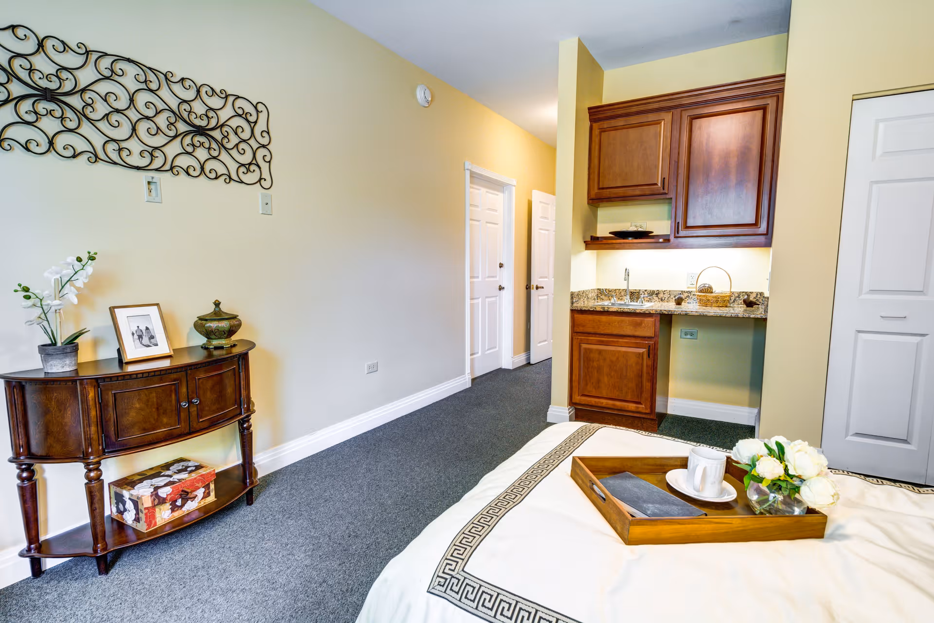 A cozy room featuring a bed with a wooden tray holding a cup, saucer, napkin, and a small vase of white flowers. To the right, there is a small kitchenette with wooden cabinets and a granite countertop. On the left side, a wooden console table holds a potted orchid, a framed photo, and a decorative jar, with a decorative wrought iron wall art above it. The room has beige walls and gray carpet flooring, with two white doors visible in the background.