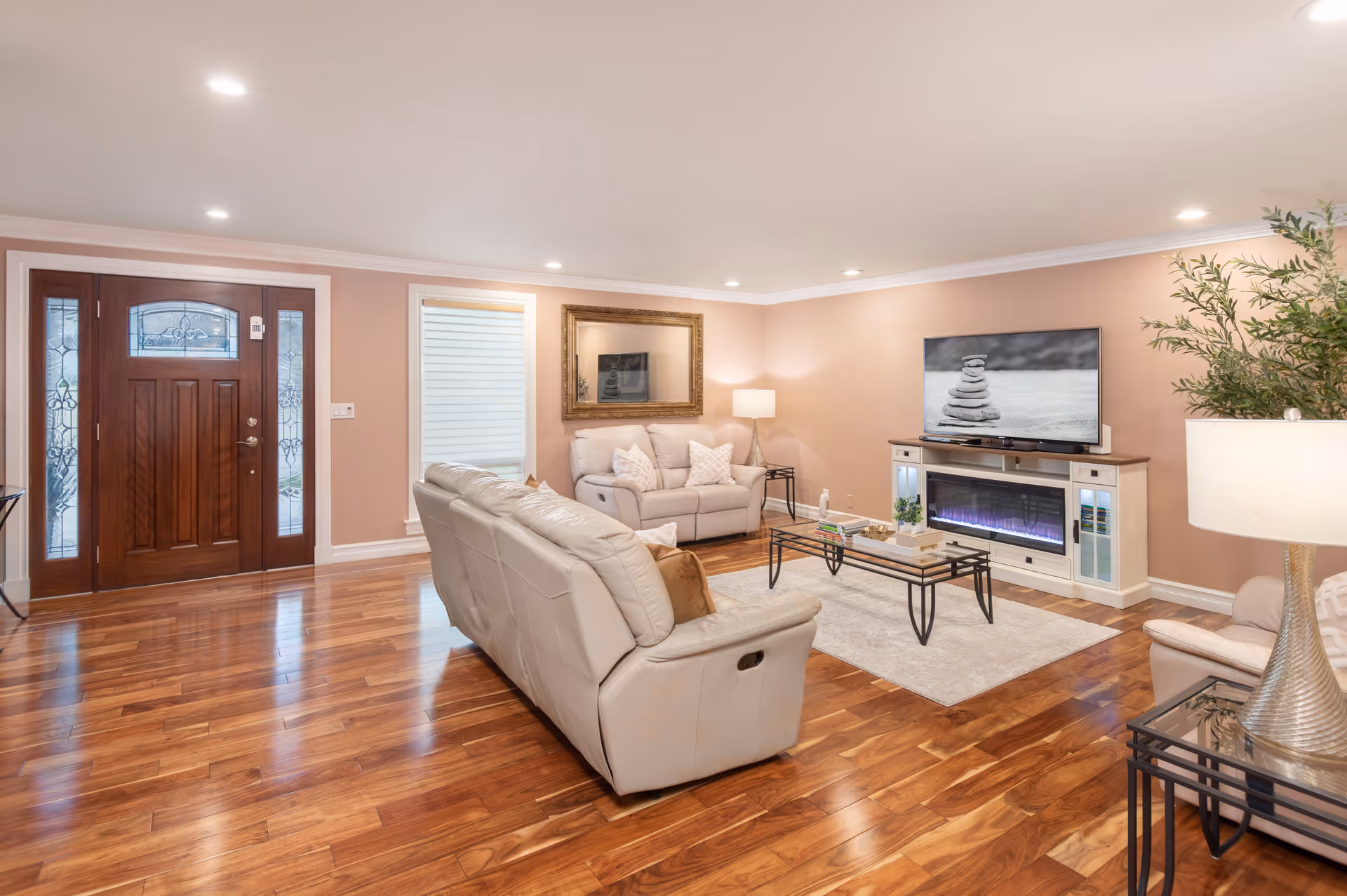 A spacious living room with polished wooden floors, beige walls, and recessed ceiling lights. The room features a wooden front door with glass panels, two beige leather sofas, a glass coffee table with decorative items, a large flat-screen TV mounted above a white electric fireplace, a floor lamp, a side table with a lamp, and a large potted plant in the corner.