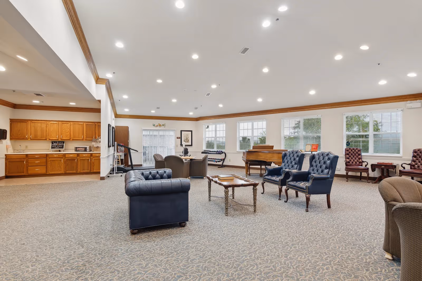 A spacious senior living facility common area with multiple seating arrangements including blue leather armchairs, brown chairs, and a wooden coffee table. The room features large windows with white blinds, a piano, and a kitchenette with wooden cabinets in the background. The ceiling has recessed lighting and crown molding along the walls.
