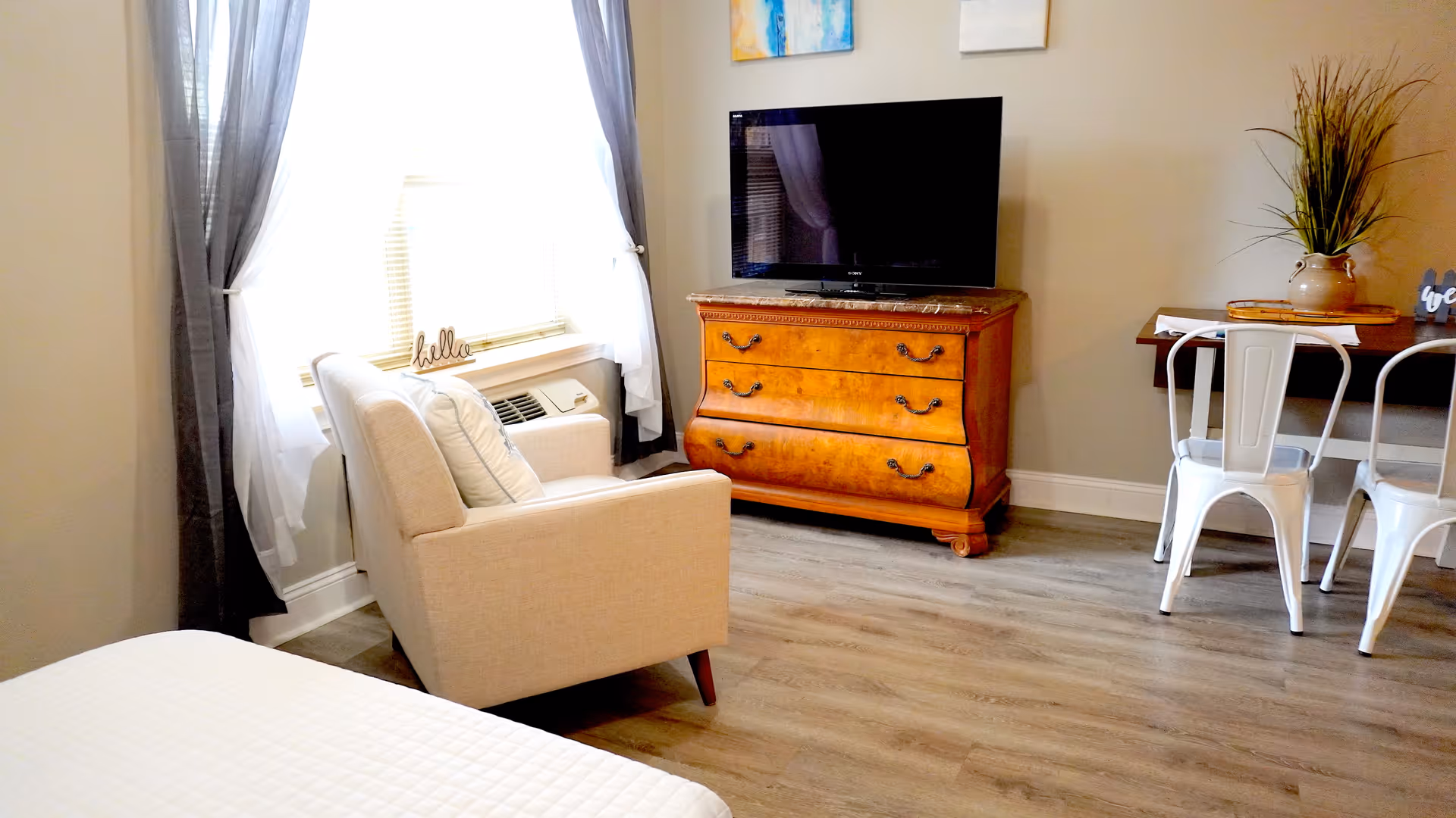 A cozy living area with a beige armchair near a window with sheer and dark curtains. A wooden dresser with a flat-screen TV on top is against the wall. To the right, there is a small dining table with white metal chairs and a decorative plant on top. The floor is light wood, and the walls are painted a soft beige.