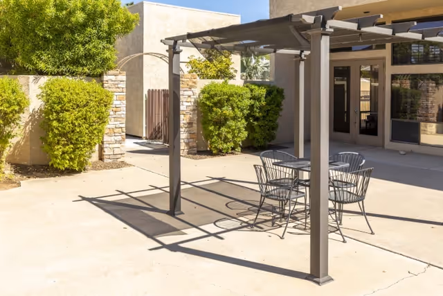 Outdoor patio area with a metal pergola casting shadows on the concrete ground, a round metal table with four matching chairs underneath, surrounded by beige walls, green bushes, and a building entrance with glass doors and windows.