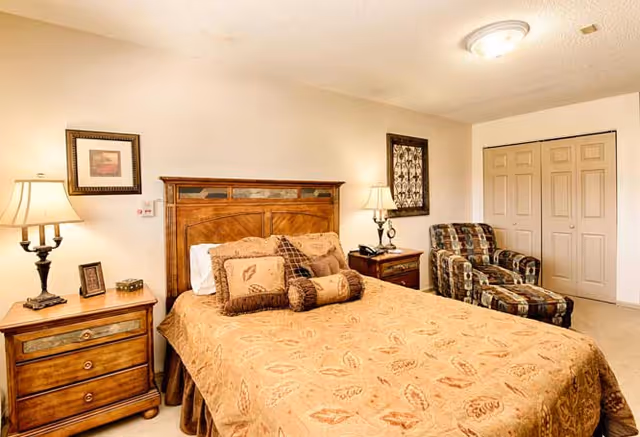 A cozy bedroom with a wooden bed frame and a bed covered in a beige patterned bedspread with multiple pillows. There are two wooden nightstands on either side of the bed, each with a lamp. A framed picture hangs on the wall above each nightstand. To the right, there is a patterned armchair with an ottoman and a closed double-door closet.