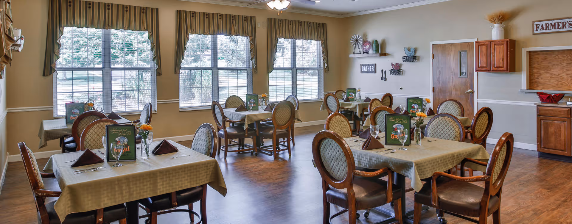 A bright dining room with multiple tables and upholstered chairs set for meals, large windows with valances, and wall decor.