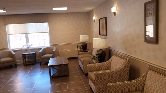 A nursing home seating area with upholstered chairs, coffee tables, lamps, and a window.